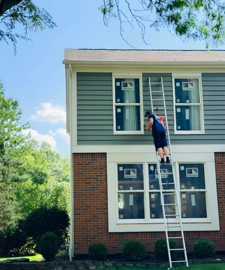 Technician on a ladder at a second-floor window of a gray-sided house with new windows