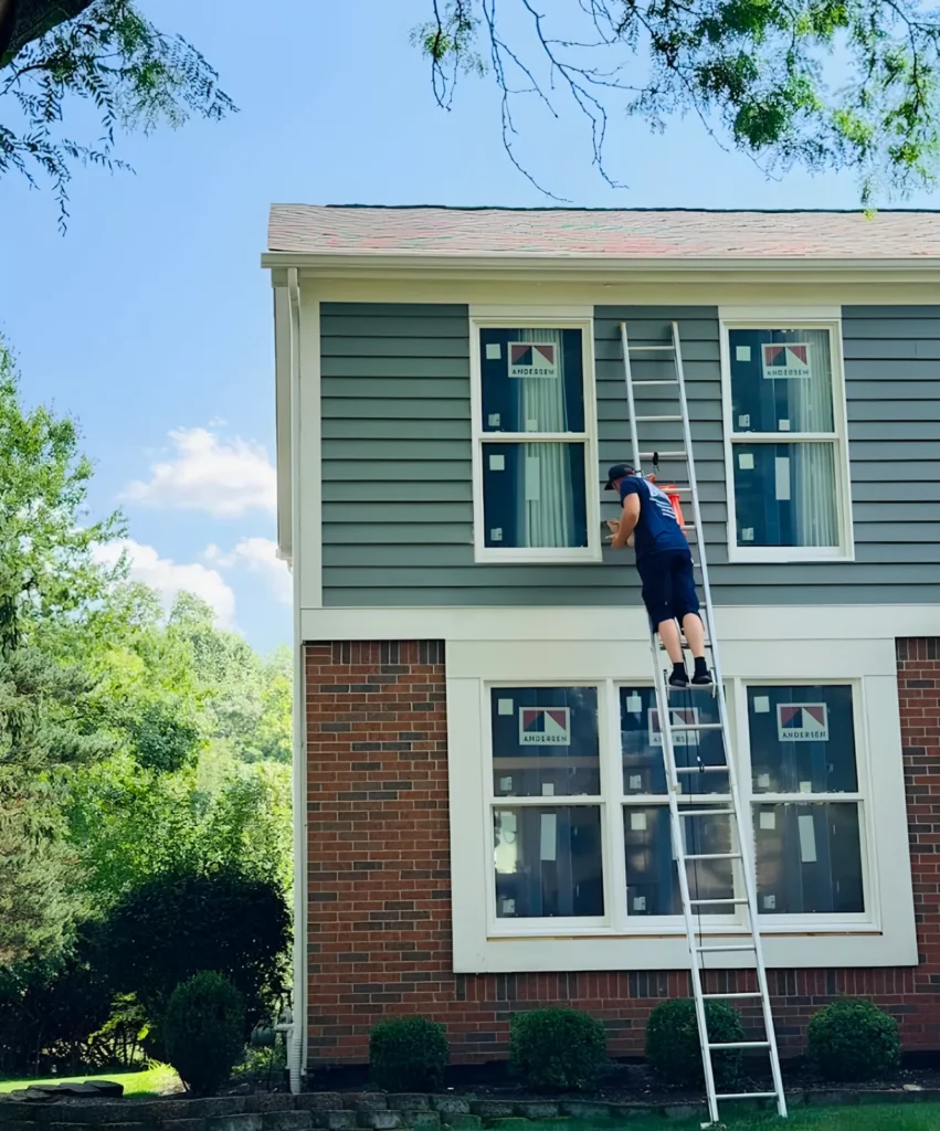 Technician on a ladder at a second-floor window of a gray-sided house with new windows