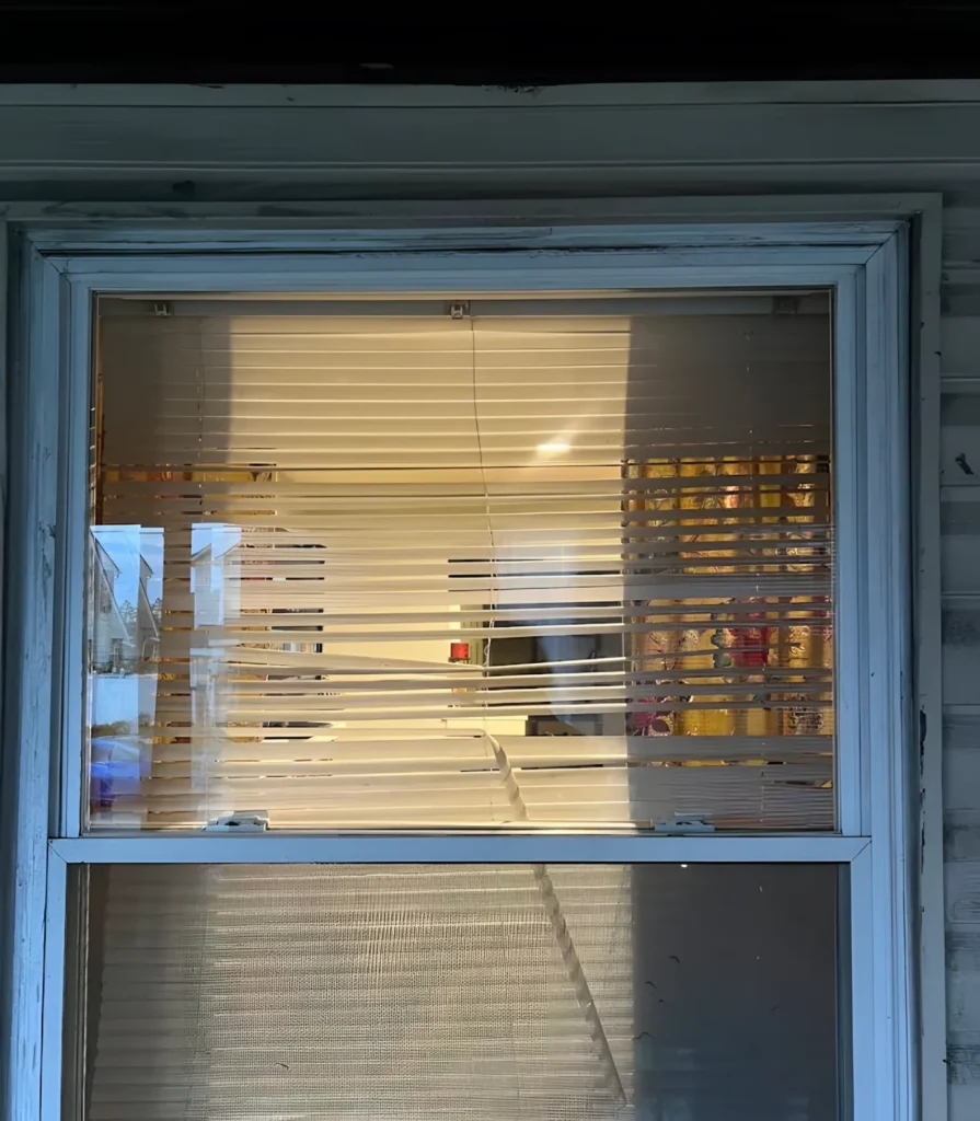 Front view of a double-hung window with a hairline crack through the upper pane and blinds behind it