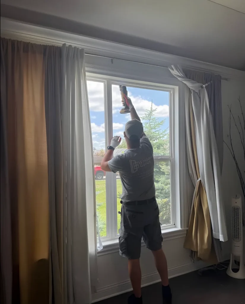 Technician in a Window Gurus shirt fitting a window with a drill, curtains on both sides