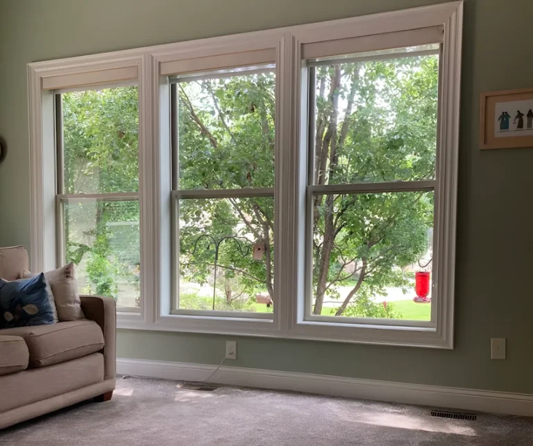 Three double-hung windows with wide white casing across a sage-green living room wall, a leafy yard and bird feeders outside