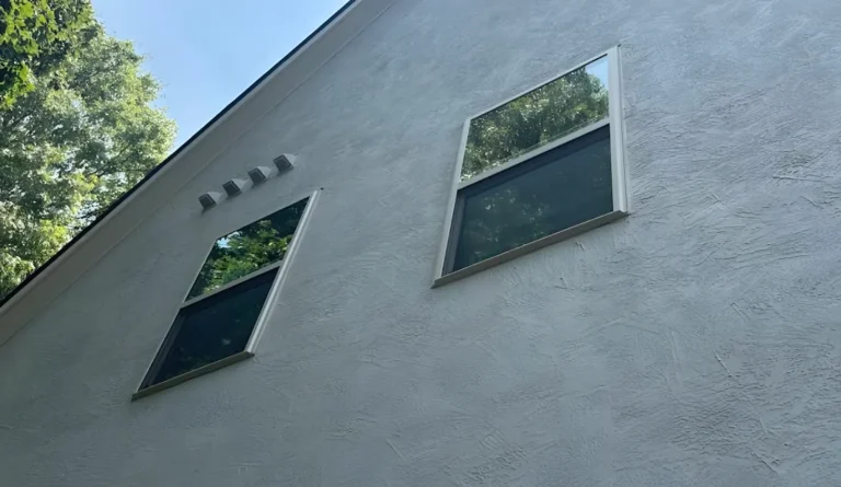 Two double-hung windows set high on a textured stucco gable wall with trees reflected in the glass, photographed from the ground looking up